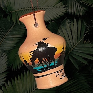 May include: Tan ceramic vase with a painted scene of a person on horseback against a sunset backdrop. The vase has a brown rim and a hanging cord. The word "Gr" is written on the vase.