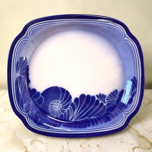 May include: A square ceramic plate with rounded corners, featuring a white center and a deep blue border. The design includes a stylized blue nautilus shell motif and leaf-like patterns along the bottom edge, framed by concentric blue lines.
