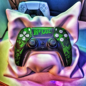 Könnte beinhalten: Ein schwarzer und grüner Videospiel-Controller mit einem grafischen Design und dem Text "JR#CUZ". Der Controller hat eine schwarze Basis mit grünen Akzenten und Tasten. Ein weiterer weißer Controller ist im Hintergrund sichtbar.