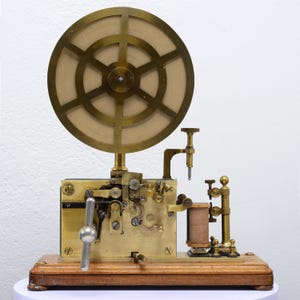 May include: Antique brass telegraph machine with a large circular wheel and intricate mechanical components. The device is mounted on a wooden base and features a lever and various other parts. A vintage communication device.