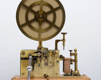 May include: Antique brass telegraph machine with a large circular wheel and intricate mechanical components. The device is mounted on a wooden base and features a lever and various other parts. A vintage communication device.