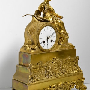 Puede incluir: Un reloj de chimenea dorado con una esfera blanca y agujas negras. El reloj presenta una figura esculpida de una mujer sentada sobre una base con intrincados detalles.