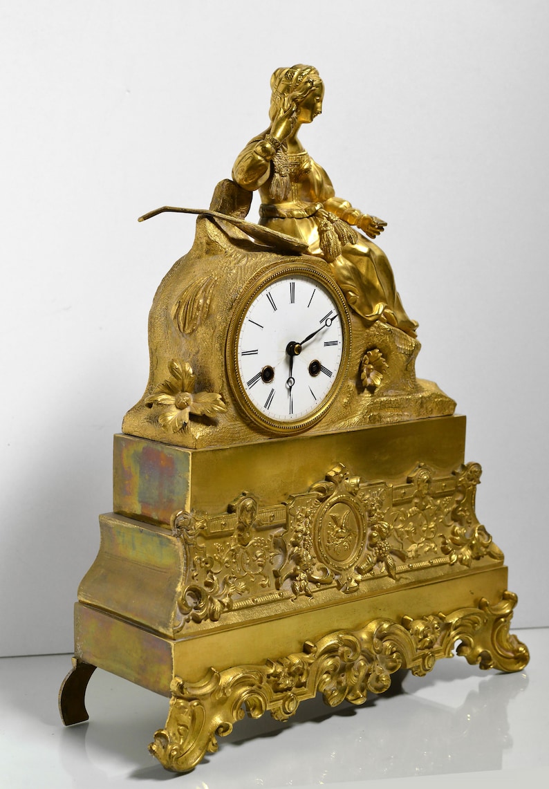 Puede incluir: Un reloj de chimenea dorado con una esfera blanca y agujas negras. El reloj presenta una figura esculpida de una mujer sentada sobre una base con intrincados detalles.