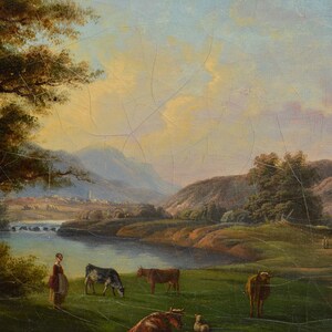 Op de afbeelding: Een schilderij dat een landelijk landschap afbeeldt met een rivier, glooiende heuvels en een klein dorp in de verte. De sc&egrave;ne is in warm, gouden licht gebaad, met een paar koeien die grazen in een weide bij de rivier. Een vrouw in een lange jurk staat aan de oever van de rivier en kijkt naar de koeien.