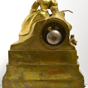Puede incluir: Un reloj de chimenea dorado con una figura femenina sentada en una roca. El reloj tiene una esfera redonda y una campana. El reloj est&aacute; sobre una base de varios niveles con un dise&ntilde;o decorativo.