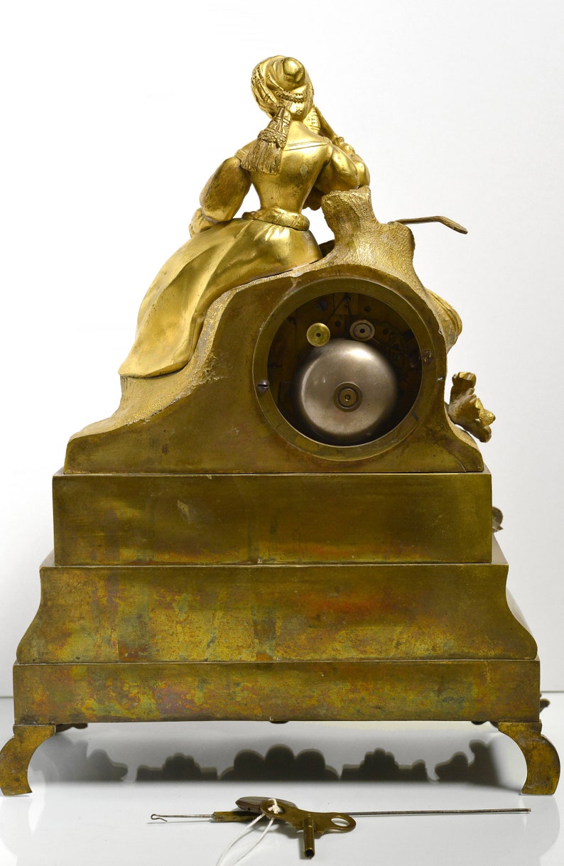 Puede incluir: Un reloj de chimenea dorado con una figura femenina sentada en una roca. El reloj tiene una esfera redonda y una campana. El reloj est&aacute; sobre una base de varios niveles con un dise&ntilde;o decorativo.