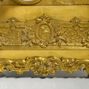 Puede incluir: Un reloj de chimenea dorado con detalles ornamentados y una base decorativa. El reloj tiene una esfera blanca con n&uacute;meros romanos negros y agujas negras.