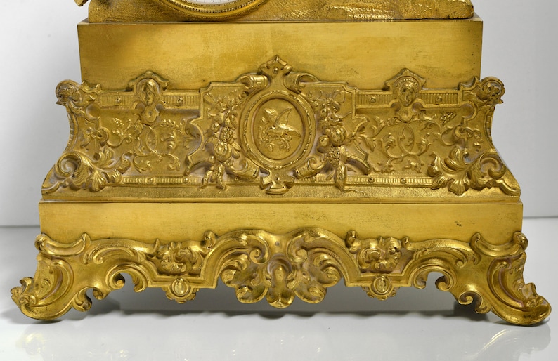 Puede incluir: Un reloj de chimenea dorado con detalles ornamentados y una base decorativa. El reloj tiene una esfera blanca con n&uacute;meros romanos negros y agujas negras.