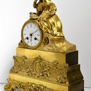 Puede incluir: Un reloj de chimenea dorado con una esfera blanca y agujas negras. El reloj presenta una figura esculpida de una mujer sentada en una roca, sosteniendo un instrumento musical. La base del reloj est&aacute; adornada con motivos florales y de volutas.