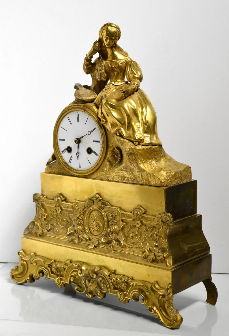 Puede incluir: Un reloj de chimenea dorado con una esfera blanca y agujas negras. El reloj presenta una figura esculpida de una mujer sentada en una roca, sosteniendo un instrumento musical. La base del reloj est&aacute; adornada con motivos florales y de volutas.