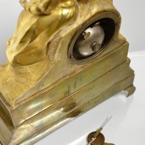 Puede incluir: Un reloj de chimenea dorado con una figura esculpida de una mujer sosteniendo un ni&ntilde;o. El reloj tiene una esfera redonda y una llave para dar cuerda.