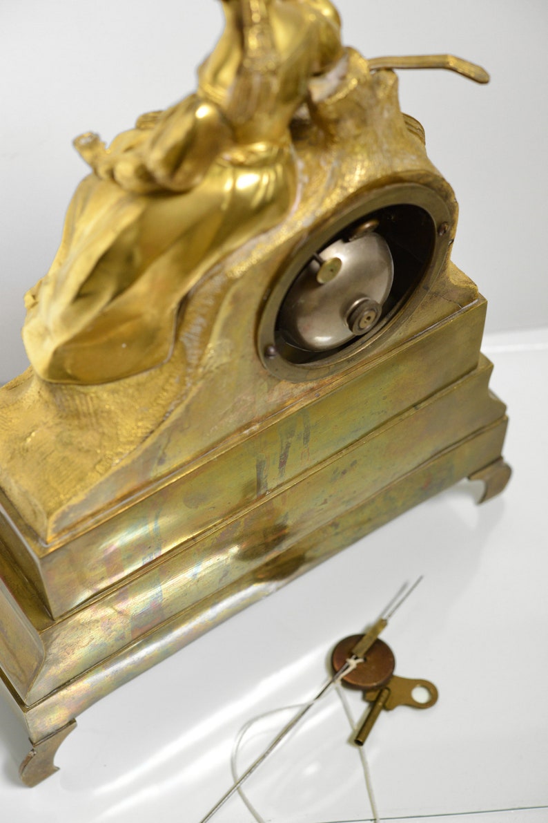 Puede incluir: Un reloj de chimenea dorado con una figura esculpida de una mujer sosteniendo un ni&ntilde;o. El reloj tiene una esfera redonda y una llave para dar cuerda.