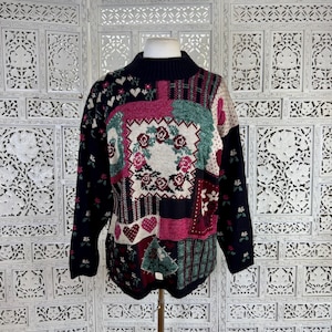 Peut inclure: Un pull à manches longues avec un col montant et un motif patchwork. Le tricot présente des sections bleu marine, crème, rose et vert avec des motifs floraux, des couronnes de roses et des cœurs sur le devant et les manches.