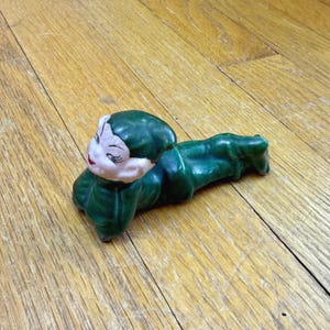以下が含まれることがあります： ダークグリーンのスーツと揃いのとんがり帽子をかぶった、横たわるエルフの陶器製フィギュア。エルフは顔色が白く、黒く描かれた目、アーチ型の眉、赤い唇が特徴です。フィギュアは腹ばいになり、頭を上げた姿勢で表現されています。