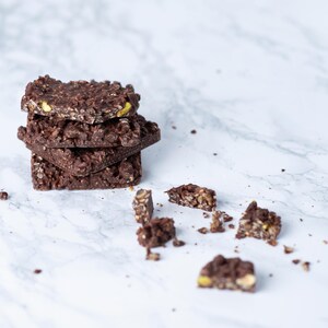May include: A stack of four dark chocolate bars with visible nuts and seeds. Some pieces are broken off and scattered around the stack. The bars are on a white marble surface.
