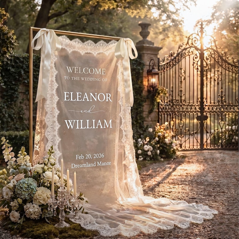 May include: A rectangular gold metal frame holds a sheer white fabric wedding sign with lace trim and satin ribbons. The text reads: WELCOME TO THE WEDDING OF ELEANOR and WILLIAM Feb 20, 2026 Dreamland Manor. A crystal candelabra with candles sits nearby.