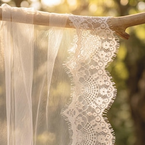 May include: A sheer, off-white veil featuring a delicate, scalloped lace trim with an intricate floral pattern and fine, fringed edges. The lightweight fabric drapes gracefully over a smooth, light-colored wooden branch, highlighting the texture.
