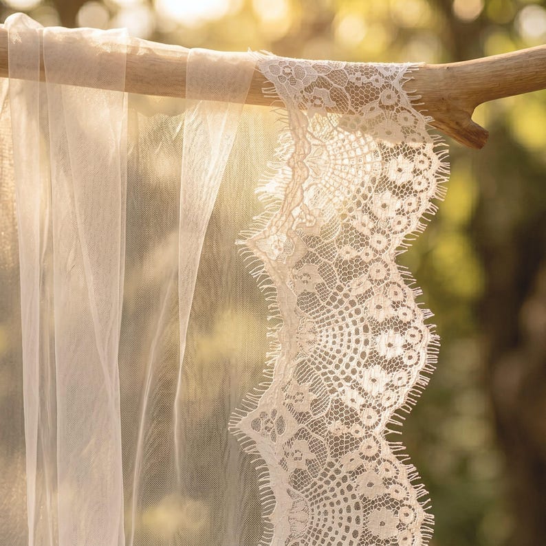 May include: A sheer, off-white veil featuring a delicate, scalloped lace trim with an intricate floral pattern and fine, fringed edges. The lightweight fabric drapes gracefully over a smooth, light-colored wooden branch, highlighting the texture.