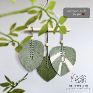 May include: Three macrame leaf wall hangings in shades of green and white, with wooden dowels and beads. The text "Customize color for you" is visible in the top right corner. The text "Macrame art by Meii" is visible on the right side of the image. The text "MeiiStudiovn meticulous in each product" is visible at the bottom of the image.