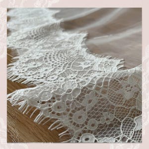 May include: A length of white lace trim featuring a scalloped edge with intricate floral patterns and a delicate mesh background. The lace has a soft texture and a repeating design that runs along the entire length of the fabric strip.