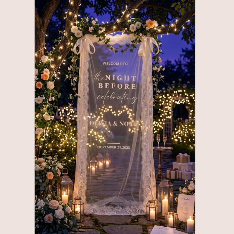 May include: A vertical sheer fabric sign with white lace trim, featuring the text "WELCOME TO the NIGHT BEFORE celebrating OLIVIA & NOAH NOVEMBER 21, 2023". The sign is suspended from a tree branch adorned with fairy lights and floral arrangements.