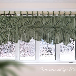 May include: Sage green macrame window valance with a leaf design. The handcrafted textile art piece is suspended from a wooden rod and hangs in front of a window overlooking a snowy landscape. The valance is labeled "Macrame art by Meii".