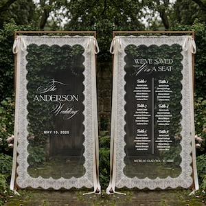 May include: Two tall rectangular wedding signs framed in wood, draped with white lace fabric and tied with ribbons. The left sign reads The Anderson Wedding May 10, 2025. The right sign lists table assignments under the text We've Saved You A Seat.