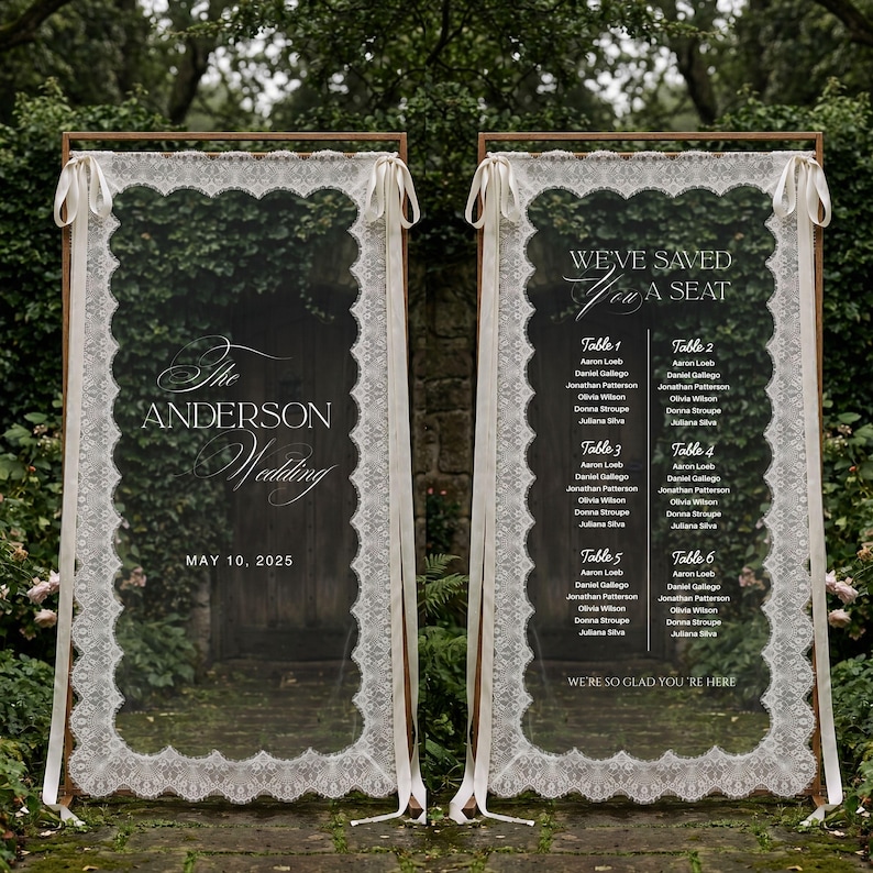 May include: Two tall rectangular wedding signs framed in wood, draped with white lace fabric and tied with ribbons. The left sign reads The Anderson Wedding May 10, 2025. The right sign lists table assignments under the text We've Saved You A Seat.
