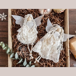 May include: Two white lace angel ornaments with silver glitter wings. The angels have white yarn hair and wooden heads. The angels are sitting on brown shredded paper.