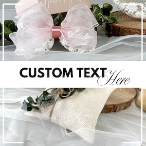 May include: A white pillow with lace trim and greenery, with two gold rings and the text "The Smiths" embroidered on it. The pillow is on a white tulle fabric with a pink bow with lace trim and the text "Addaline" on it.