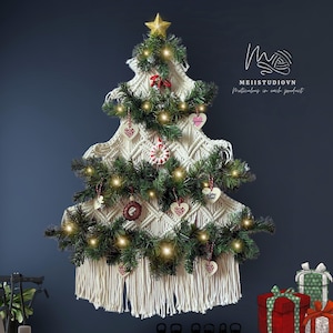 May include: A macrame Christmas tree with a gold star topper, decorated with faux evergreen branches, heart-shaped ornaments, and string lights. The tree has a fringed bottom and is displayed against a dark blue background.