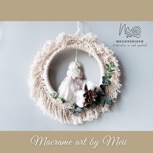 May include: A white macrame wreath with a white gnome figurine, cotton balls, and eucalyptus leaves. The wreath is hanging from a white wall. The text "Macrame art by Meii" is at the bottom of the image.