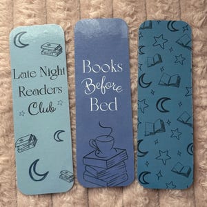 May include: Three rectangular bookmarks with rounded corners. One is light blue with the text "Late Night Readers Club" and illustrations of books and moons. The second is blue with "Books Before Bed" and a coffee cup. The third is blue with book, moon, and star patterns.
