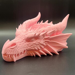 Peut inclure: Une sculpture de tête de dragon rose clair présentant des textures d'écailles complexes, des pointes faciales acérées et des cornes proéminentes. La créature a la bouche fermée avec des dents visibles et une section supérieure creuse.
