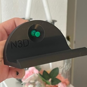 Può includere: Un supporto per telefono nero a forma di semicerchio con le lettere "N3D" in rilievo sulla parte anteriore. È dotato di un piccolo pulsante verde e di un ripiano per tenere un telefono. Il supporto è tenuto in mano, con uno sfondo sfocato di candele e fiori.