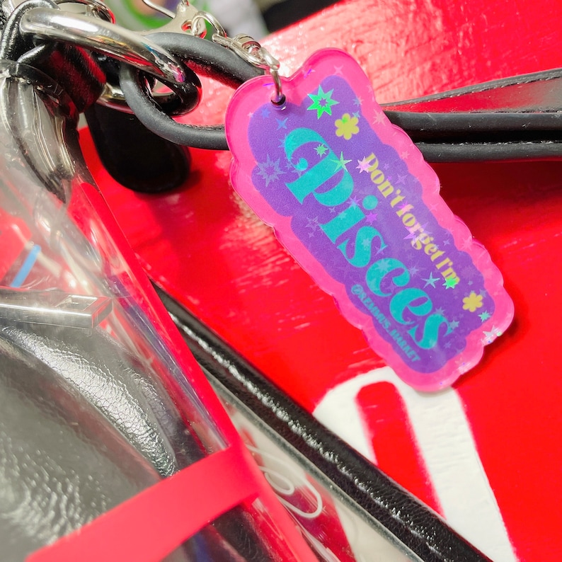 Puede incluir: Un llavero hologr&aacute;fico rosa y morado con el texto "No olvides que soy Piscis" y estrellas. El llavero est&aacute; unido a una correa negra.