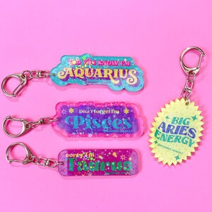 Puede incluir: Cuatro llaveros coloridos con diferentes signos del zod&iacute;aco: Acuario, Piscis, Aries y Tauro. Cada llavero presenta un fondo brillante con estrellas y purpurina.