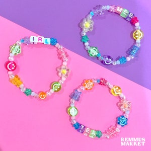 May include: Three colorful beaded bracelets with different words spelled out on them. The bracelets are made with clear beads, rainbow colored beads, and smiley face beads. The words on the bracelets are "IRL", "MEEE", and "TEA".