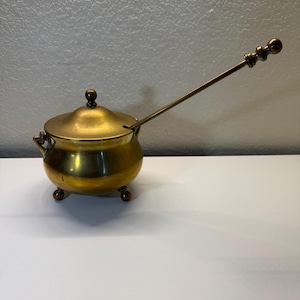 May include: A brass pot with a rounded body, three small feet, and two side handles. It features a removable lid with a central knob and a long, slender brass spoon resting inside, extending outward from a small notch in the lid rim.