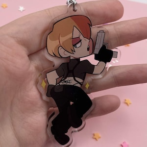 Op de afbeelding: Een doorzichtige acryl sleutelhanger met een cartoonfiguur met tweekleurig haar, gekleed in een zwarte outfit met een harnas en een grijs handvuurwapen vasthoudend. Het figuur heeft een tribale tatoeage op de onderrug en een dynamische houding.