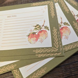 Peut inclure: Une pile de fiches de recettes rectangulaires avec un centre crème, des lignes horizontales et le mot DIRECTIONS imprimé en brun. Les cartes présentent une bordure vert olive avec des motifs de feuilles et une illustration aquarelle de deux pêches.