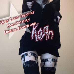 Op de afbeelding: Zwarte gehaakte tas met het Korn-logo in rood en wit. De tas wordt vastgehouden door een persoon die zwarte kleding draagt. De tekst op de afbeelding luidt "Korn Logo Crochet Bag Pattern Digital Download". De tas heeft een rechthoekige vorm.