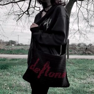 May include: Black crochet shoulder bag with the word "Deftones" in red lettering. The bag is worn over the shoulder. The person in the image is wearing a black hoodie and black pants. The background includes a tree and grass.