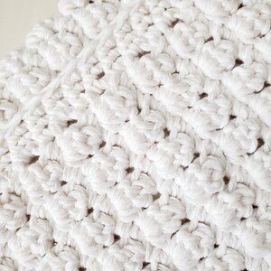 May include: A white crocheted textile featuring a repeating textured pattern of raised clusters. The fabric is composed of thick, soft yarn loops arranged in diagonal rows, creating a dense, uniform surface with a consistent matte finish.