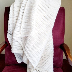 May include: A white crocheted blanket draped over a burgundy armchair. The blanket is folded and has a textured, woven pattern.