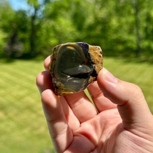 May include: A polished opal gemstone with a dark, iridescent interior and a rough, tan exterior. The opal displays flashes of green, blue, and gold. The stone is held in a hand against a blurred green background.