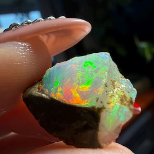 Op de afbeelding: Een ruwe opaal edelsteen met een levendige weergave van kleuren, waaronder groen, rood en geel. De opaal wordt tussen vingers gehouden, wat de natuurlijke, onbewerkte textuur en het iriserende kleurenspel benadrukt. De steen heeft een donkerbruine basis.