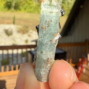 Op de afbeelding: Een doorschijnend, lichtblauw-groen kristal met een rechthoekige vorm. Het kristal heeft een enigszins ruwe textuur en wordt in een hand gehouden. Kleine, donkere minerale afzettingen zijn aan het kristal bevestigd.