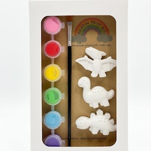 May include: A white cardboard box containing a craft kit with three white plaster dinosaur figures, a black paintbrush, and a strip of six small paint pots in pink, red, yellow, green, blue, and purple. The packaging features a rainbow logo and text.