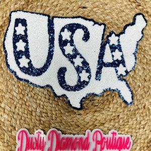 May include: A white and blue glitter USA map-shaped decoration. The word "USA" is spelled out in blue glitter, with white star accents. The decoration is on a woven background, with a pink and white "Dusty Diamond Boutique" logo below.